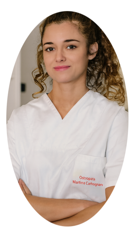 Osteopata Martina Carbognani D.O. Una professionista sanitario Osteopata in camice bianco sta in piedi con le braccia incrociate in un ufficio medico illuminato dalla luce naturale, con una sedia ergonomica, un tavolo, un computer e uno scheletro anatomico visibili sullo sfondo