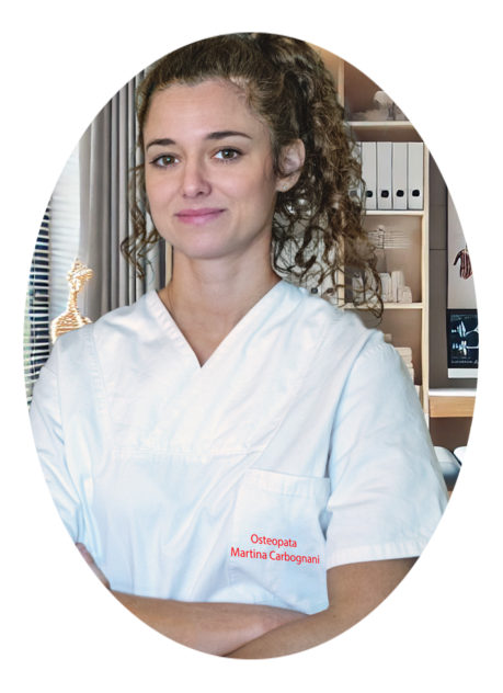 Una professionista sanitario Osteopata in camice bianco sta in piedi con le braccia incrociate in un ufficio medico illuminato dalla luce naturale, con una sedia ergonomica, un tavolo, un computer e uno scheletro anatomico visibili sullo sfondo