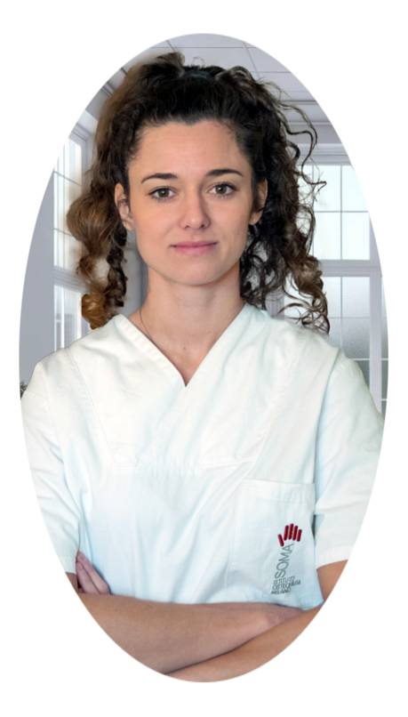 Osteopata Martina Carbognani D.O. Una professionista sanitario Osteopata in camice bianco sta in piedi con le braccia incrociate in un ufficio medico illuminato dalla luce naturale, con una sedia ergonomica, un tavolo, un computer e uno scheletro anatomico visibili sullo sfondo